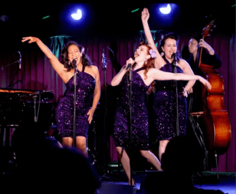 Three women in sequined purple dresses singing on stage with a double bass player in Central Oregon.