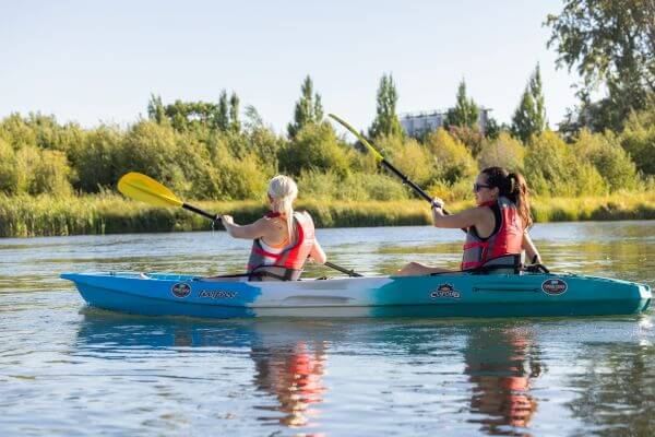 Two women paddling a tandem kayak on a calm river near green trees in Central Oregon.