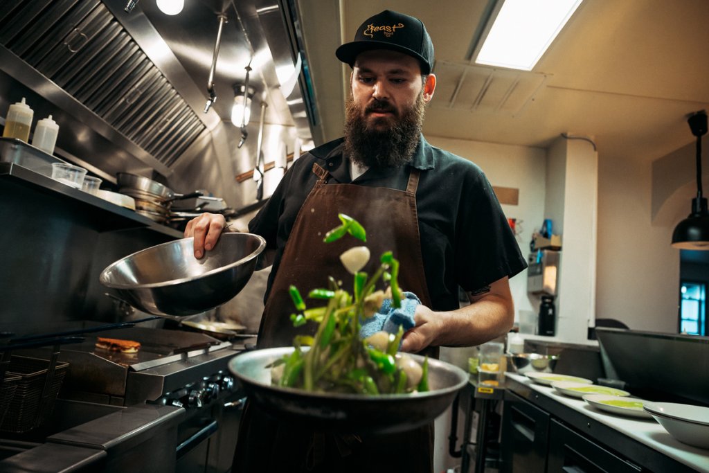A bearded chef tossing green vegetables in a pan inside a professional kitchen in Central Oregon.