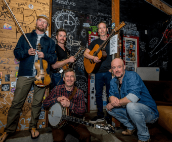 A four-piece folk band posing with musical instruments in a graffiti-covered room in Central Oregon.