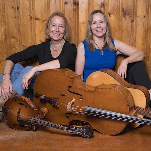 Two smiling women posing with a cello, guitar, and mandolin against a wooden wall in Central Oregon.