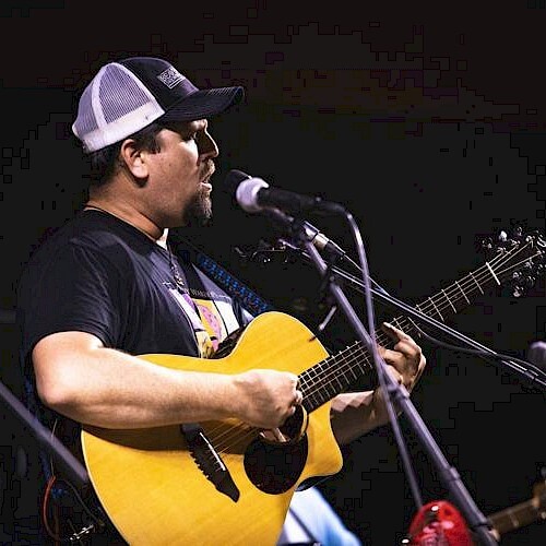 A man in a baseball cap playing an acoustic guitar and singing into a microphone in Central Oregon.