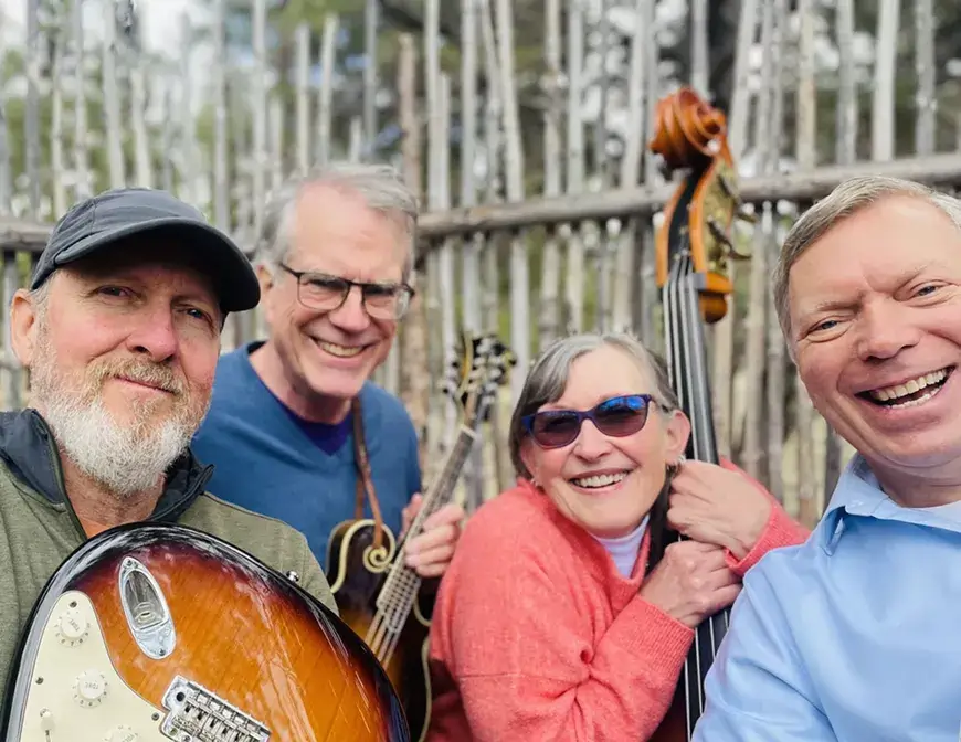 Four smiling musicians posing together with their instruments in front of a wooden fence in Central Oregon.