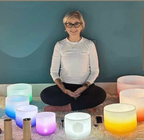 A woman sits cross-legged surrounded by glowing, colorful crystal singing bowls in Central Oregon.