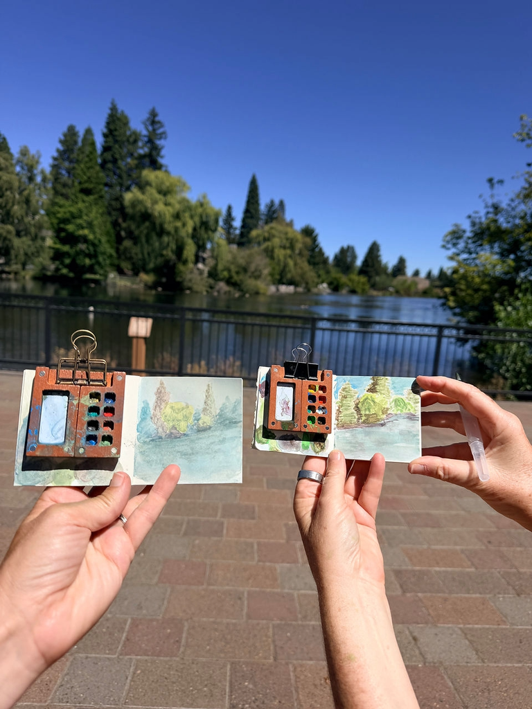 Two hands hold small watercolor sketchbooks with paint palettes in front of a lake scene in Central Oregon.