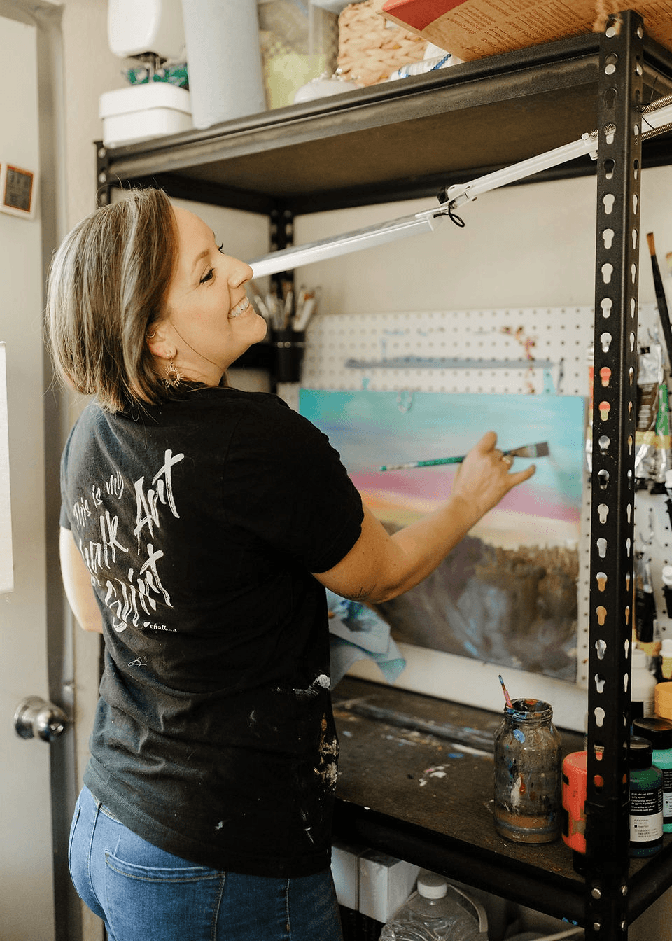A smiling woman paints on a canvas in her art studio in Central Oregon.