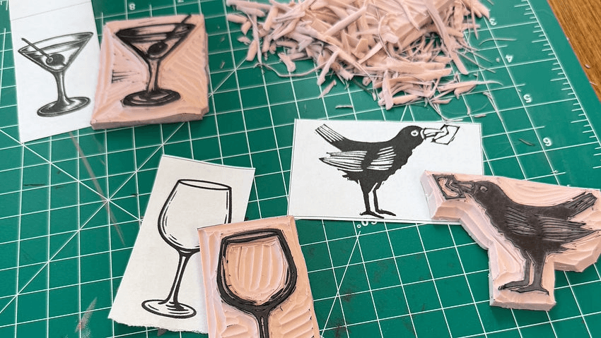 Linocut stamps of a martini glass and a crow alongside their printed paper proofs in Central Oregon.