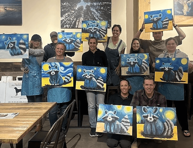 A group of people proudly displaying their raccoon paintings at an art class in Central Oregon.