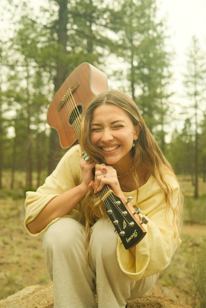 A smiling young woman crouching in a forest holding an acoustic guitar in Central Oregon.