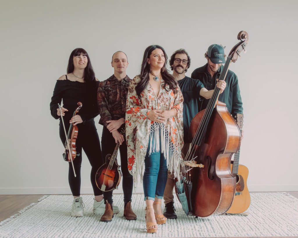 A five-piece folk band posing together with their musical instruments against a plain wall in Central Oregon.