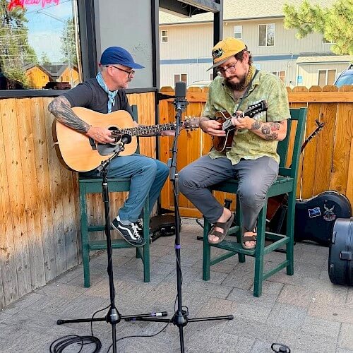 Two men playing acoustic guitar and mandolin while sitting on stools outdoors in Central Oregon.