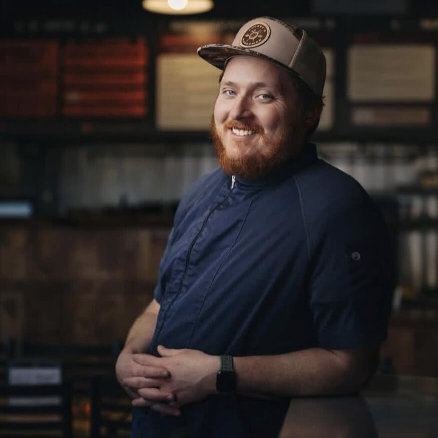 A smiling man with a red beard wearing a baseball cap and chef's uniform in Central Oregon.