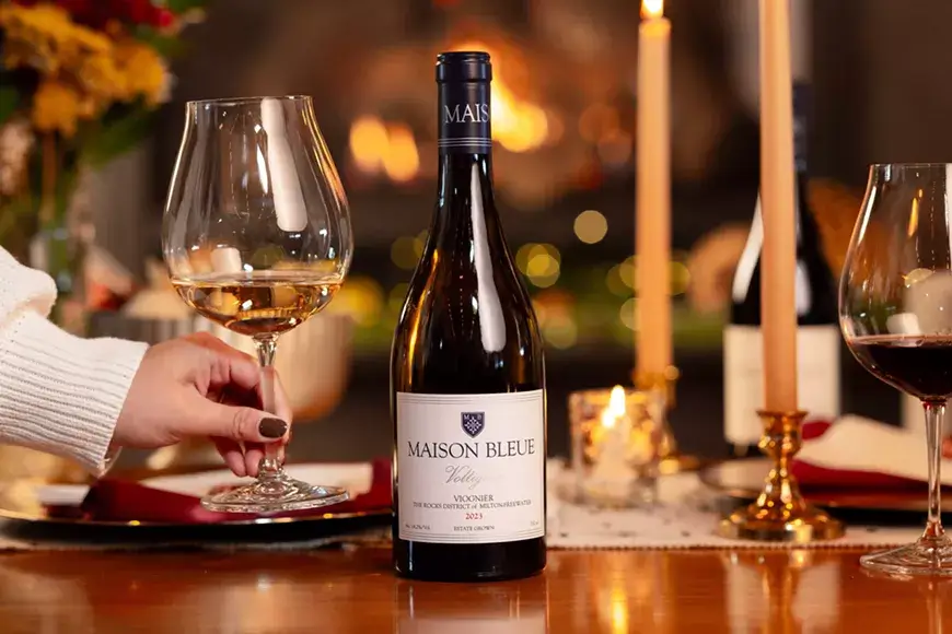A hand holds a glass of white wine beside a bottle of Maison Bleue in Central Oregon.